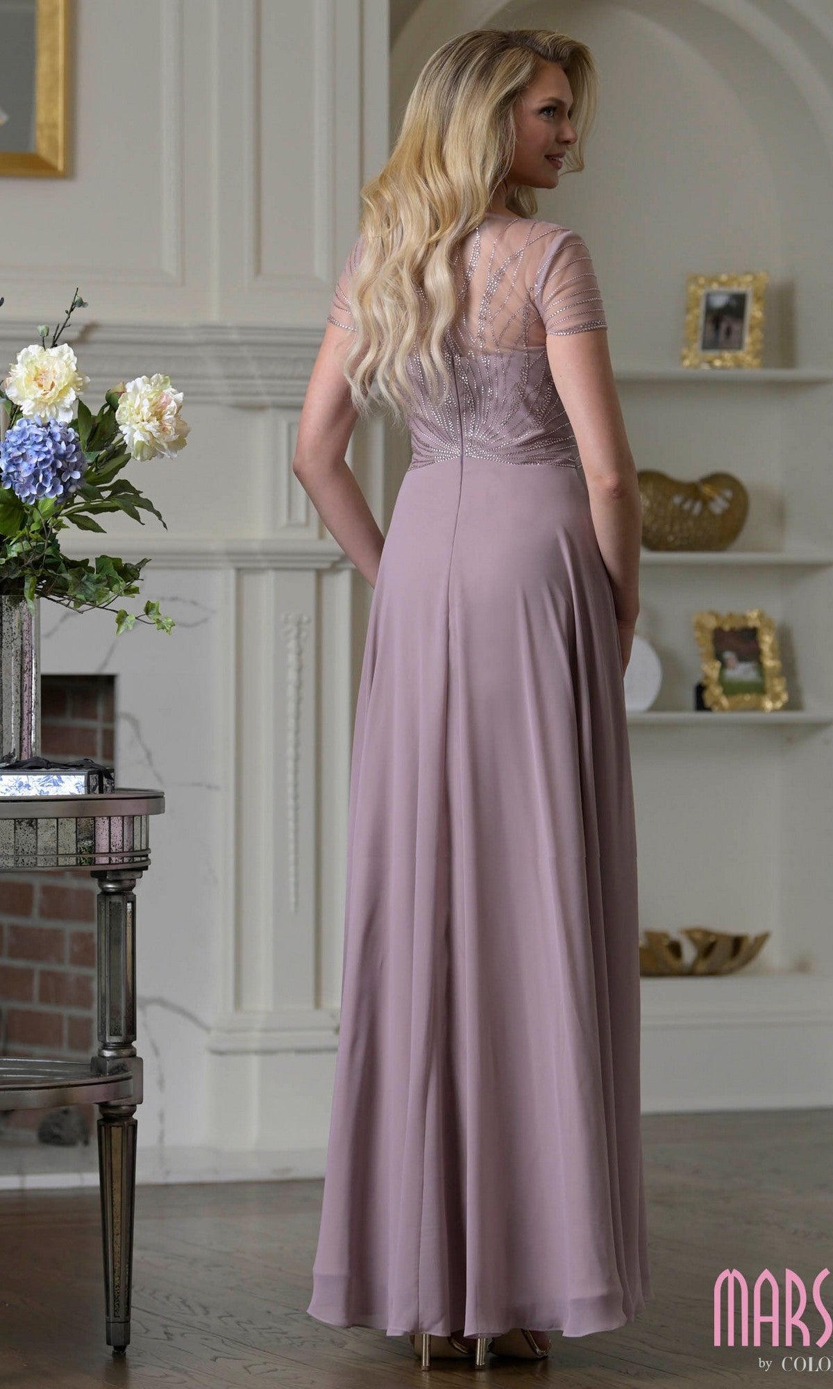 A woman with long blonde hair stands indoors, facing away from the camera, wearing the Marsoni M351 long formal mauve chiffon A-line gown with sheer short sleeves.